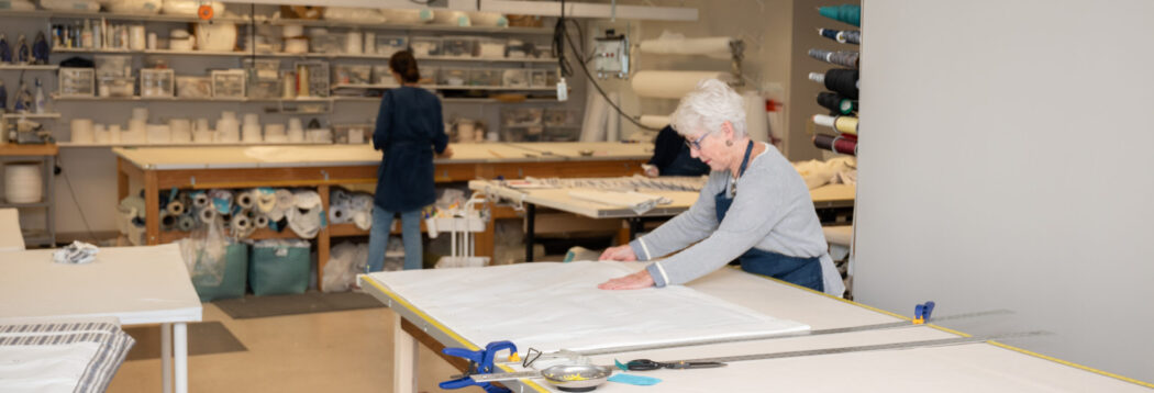 Two workroom staff laying out and cutting drapery fabric at long tables, with rolls of fabric shelved behind them.