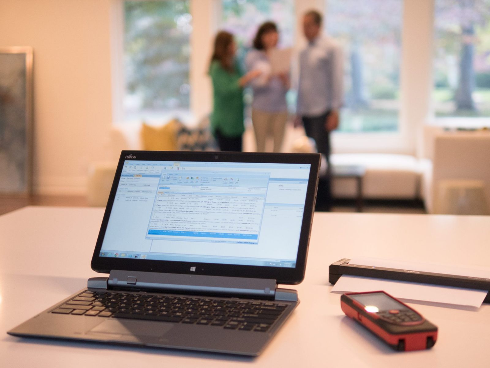 A laptop running Solatech SalesPRO on a table beside a Leica laser distance meter and a portable printer, with a salesperson and two clients talking in the room behind.